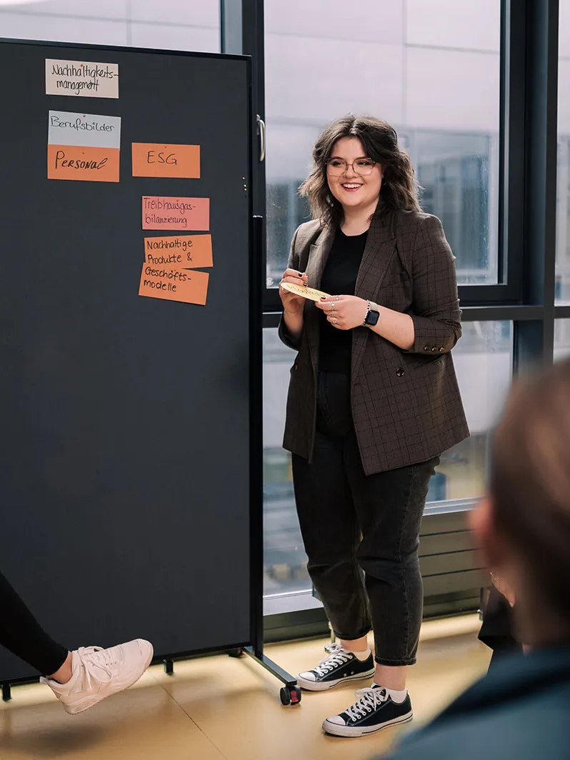 Eine Studentin steht vor einer Pinwand und erklärt Begriffe aus dem Nachhaltigkeitsmanagement.